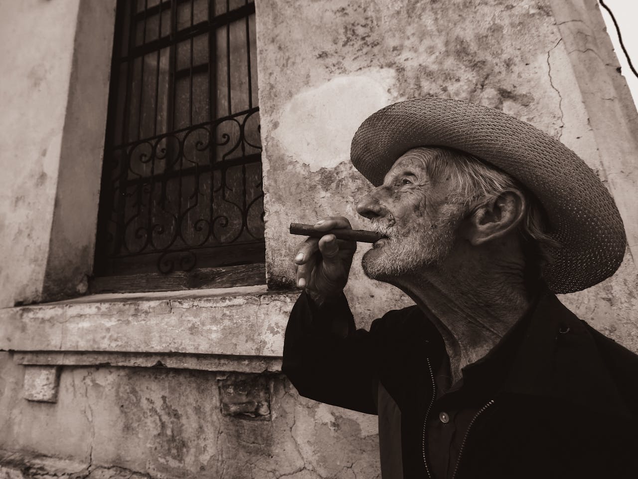 An elderly man with a cigar, wearing a straw hat, in a sepia-toned urban environment.